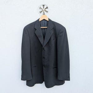 May include: A dark gray blazer with a notched lapel, three-button closure, and two front pockets. The blazer is hanging on a wooden hanger against a white wall. The blazer is a classic style.