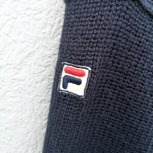 Puede incluir: Primer plano de un su&eacute;ter de punto azul marino. El su&eacute;ter presenta una textura acanalada y un peque&ntilde;o parche bordado con un logotipo rojo, blanco y azul en la manga. La tela parece suave y c&aacute;lida.