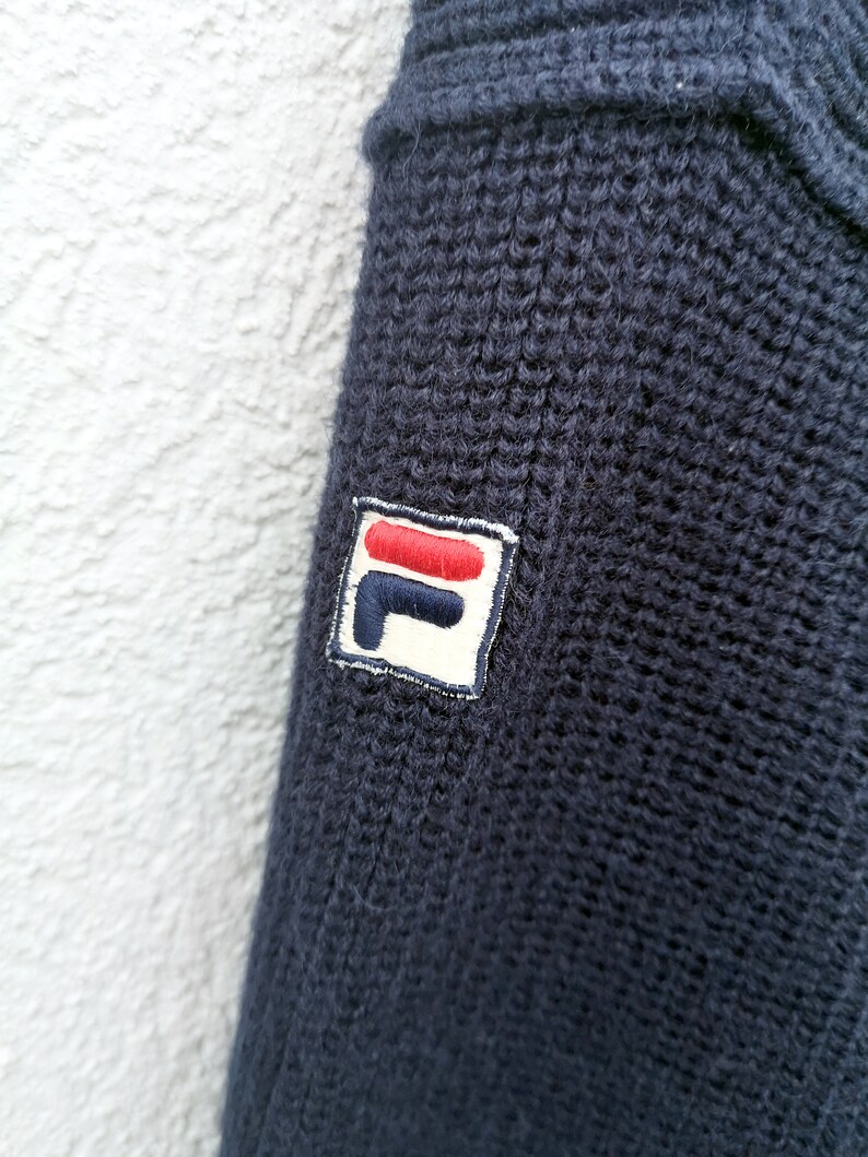 Puede incluir: Primer plano de un su&eacute;ter de punto azul marino. El su&eacute;ter presenta una textura acanalada y un peque&ntilde;o parche bordado con un logotipo rojo, blanco y azul en la manga. La tela parece suave y c&aacute;lida.