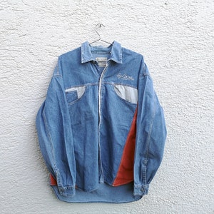 May include: A blue denim shirt with a red and gray panel on the right side. The shirt has a zipper closure and a small embroidered logo that says "By Boon".