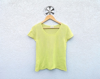 May include: A short-sleeved, lime green knit top hangs on a wooden hanger. The shirt has a scoop neckline and a slightly textured appearance. The background is a textured white wall with a metal vent.