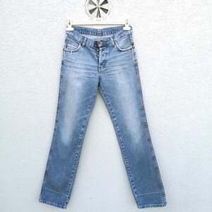 May include: Light blue denim jeans with a button fly and five pockets. The jeans are hanging on a white hanger against a white wall.