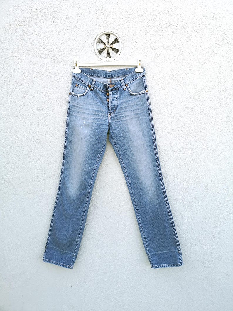 May include: Light blue denim jeans with a button fly and five pockets. The jeans are hanging on a white hanger against a white wall.
