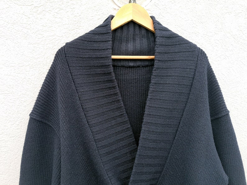 Puede incluir: Un c&aacute;rdigan azul marino oscuro con cuello chal. El su&eacute;ter est&aacute; hecho de una tela de punto acanalado y cuelga de una percha de madera. La prenda tiene un ajuste relajado y un dise&ntilde;o cl&aacute;sico.