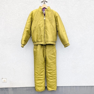 May include: A vintage gold-colored ski suit, featuring a quilted jacket with a zip closure and matching pants. The jacket has a purple lining and ribbed cuffs. The suit is displayed against a white wall.
