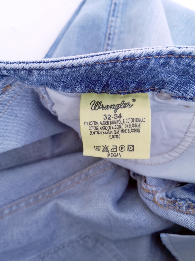 May include: A light blue denim jean label with the Wrangler logo and the text "32-34" and care instructions. The label also includes the text "97% Cotton, 3% Elastane".