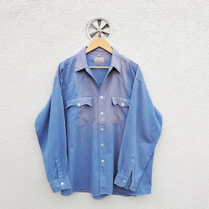 Può includere: Una camicia a maniche lunghe di colore azzurro chiaro con bottoni, colletto a punta e due tasche sul petto. La camicia ha un aspetto sbiadito ed è appesa a un appendiabiti di legno.