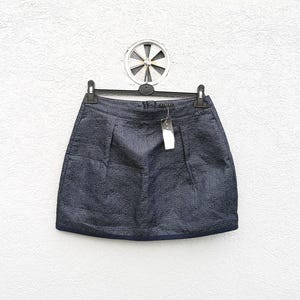 May include: A dark navy blue mini skirt with a subtle textured weave pattern. The skirt features a structured waistband, two vertical pleats on the front, and a small white rectangular tag attached to the waistband. It is displayed on a black hanger.