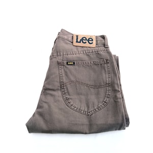 May include: Folded pair of brown Lee jeans with a leather patch on the back waistband. The jeans have a classic five-pocket design and a dark brown wash. The Lee logo is visible on the patch and the back pocket.