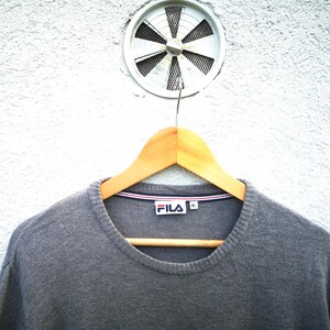 May include: A grey crew neck sweater with the Fila logo on the chest, hanging on a wooden hanger. The sweater has a ribbed neckline with red, white, and blue trim. The background is a white textured wall with a circular vent.