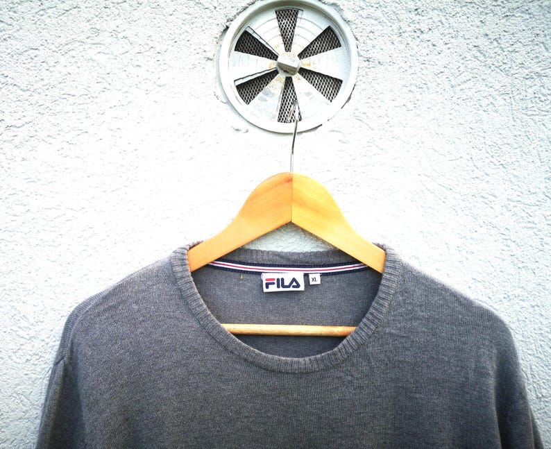 May include: A grey crew neck sweater with the Fila logo on the chest, hanging on a wooden hanger. The sweater has a ribbed neckline with red, white, and blue trim. The background is a white textured wall with a circular vent.