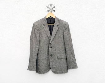 May include: A gray tweed blazer with a two-button closure, hanging on a wooden hanger. The jacket has a notched lapel, two front pockets, and a dark navy blue lining. The blazer is displayed against a white textured wall.