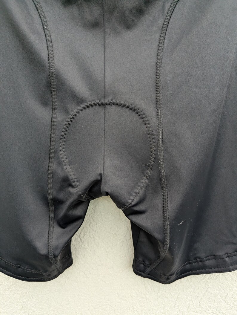 Puede incluir: Pantalones cortos de ciclismo negros con una inserci&oacute;n acolchada central con costuras en zigzag visibles a lo largo de los bordes. La prenda est&aacute; confeccionada con un tejido liso y el&aacute;stico con costuras reforzadas que recorren verticalmente los paneles frontales.