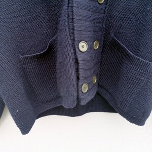Puede incluir: Primer plano de un c&aacute;rdigan azul marino. El su&eacute;ter tiene dos bolsillos delanteros y se abrocha con cuatro botones redondos de color claro. El tejido de punto es un tejido grueso y texturizado.