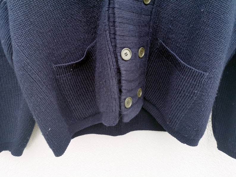 Puede incluir: Primer plano de un c&aacute;rdigan azul marino. El su&eacute;ter tiene dos bolsillos delanteros y se abrocha con cuatro botones redondos de color claro. El tejido de punto es un tejido grueso y texturizado.