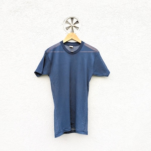 Puede incluir: Una camiseta de malla azul marino cuelga de una percha de madera contra una pared blanca texturizada. La camiseta de manga corta tiene cuello redondo y un aspecto semitransparente. La parte superior es de color liso, con un tono más oscuro alrededor del cuello.