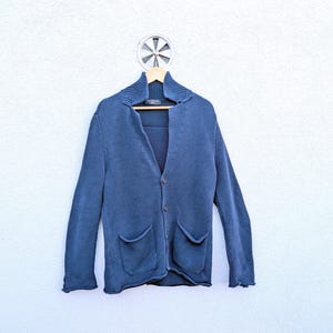 Puede incluir: Un cárdigan de punto azul con cuello de chal acanalado, mangas largas, dos bolsillos delanteros de parche y cierre frontal de botones. La prenda se muestra en una percha de madera sobre una pared de color claro con textura.