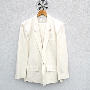 Op de afbeelding: Een wit blazer met een enkele knoopsluiting en twee voorzakken. De blazer hangt aan een houten hanger tegen een witte muur.
