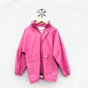 Puede incluir: Chaqueta rosa brillante con capucha y cierre de cremallera, colgada de una percha de madera. La chaqueta tiene bolsillos y una capucha con cordón. El fondo es una pared blanca texturizada con una ventilación circular.