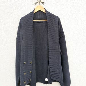 Puede incluir: Un c&aacute;rdigan azul marino con cuello chal y cierre de botones cruzados. El su&eacute;ter est&aacute; colgado en una percha de madera contra una pared blanca. Los botones son de color lat&oacute;n.