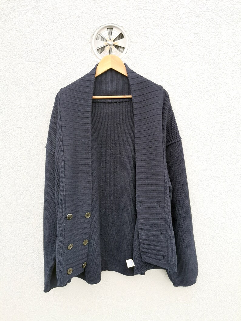 Puede incluir: Un c&aacute;rdigan azul marino con cuello chal y cierre de botones cruzados. El su&eacute;ter est&aacute; colgado en una percha de madera contra una pared blanca. Los botones son de color lat&oacute;n.