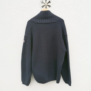 Puede incluir: Un su&eacute;ter azul marino con cuello chal, colgado de una percha de madera. La parte trasera del su&eacute;ter es visible, mostrando un patr&oacute;n de punto texturizado. Un peque&ntilde;o logotipo est&aacute; en la manga izquierda. El su&eacute;ter se exhibe contra una pared blanca.