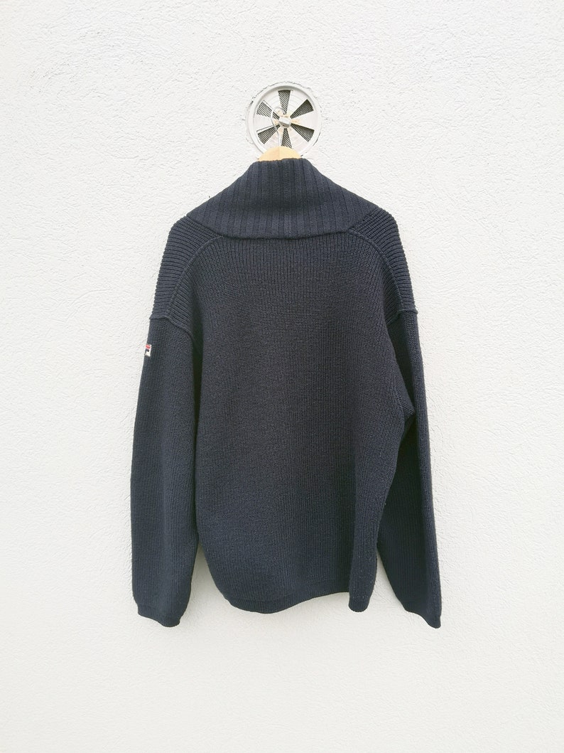 Puede incluir: Un su&eacute;ter azul marino con cuello chal, colgado de una percha de madera. La parte trasera del su&eacute;ter es visible, mostrando un patr&oacute;n de punto texturizado. Un peque&ntilde;o logotipo est&aacute; en la manga izquierda. El su&eacute;ter se exhibe contra una pared blanca.