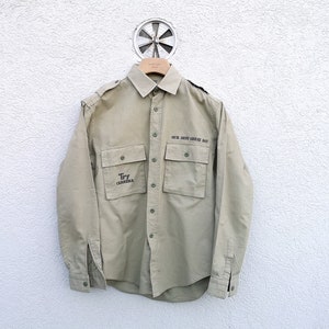 May include: Khaki-colored button-down shirt with two chest pockets and epaulets. The shirt features the text "OUR NEXT GREAT DAY" above the right pocket and "Try CARRERA" on the left pocket. The shirt is hanging on a wooden hanger.