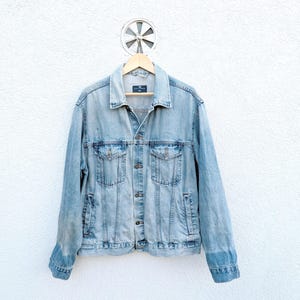 May include: A light-wash blue denim jacket featuring a classic collar, a button-up front, two chest pockets with buttoned flaps, and long sleeves with buttoned cuffs. The jacket is displayed on a wooden hanger against a textured white wall.