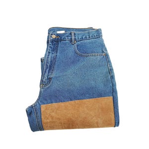 Puede incluir: Vaqueros azules con un parche de ante marrón claro en la rodilla. Los vaqueros tienen un diseño clásico de cinco bolsillos y cierre de botón y cremallera. El denim es de lavado medio.
