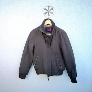 Può includere: Una giacca bomber grigia scura con chiusura a zip e due tasche. La giacca ha una fodera a quadri bianco e nero visibile al collo.