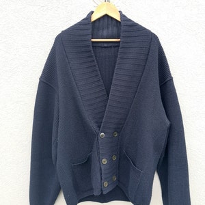 Puede incluir: Un c&aacute;rdigan azul marino con cuello chal y cierre de botones cruzados. El su&eacute;ter tiene dos bolsillos delanteros y est&aacute; hecho de un tejido de punto grueso. Cuelga de una percha de madera contra una pared blanca texturizada.