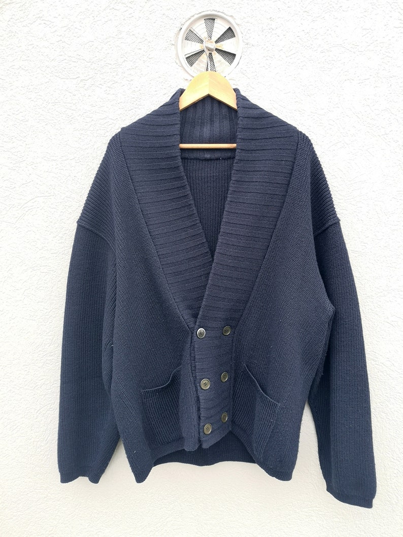 Puede incluir: Un c&aacute;rdigan azul marino con cuello chal y cierre de botones cruzados. El su&eacute;ter tiene dos bolsillos delanteros y est&aacute; hecho de un tejido de punto grueso. Cuelga de una percha de madera contra una pared blanca texturizada.