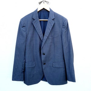 May include: A navy blue blazer with a subtle pinstripe pattern, hanging on a wooden hanger. The blazer has two front pockets and a two-button closure. The background is a plain white wall.