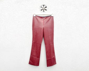 Può includere: Un paio di pantaloni in similpelle rosso scuro con finitura liscia semimatta, taglio a gamba dritta e chiusura centrale con zip sul davanti. I pantaloni sono esposti su una gruccia di plastica bianca contro una parete bianca testurizzata.