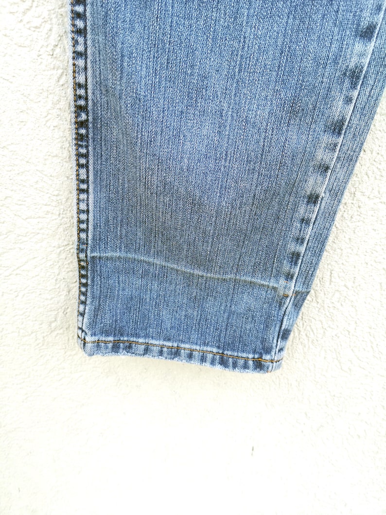 May include: Close-up of the bottom hem of a pair of blue denim jeans. The hem is folded and stitched with a dark thread.