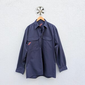 Puede incluir: Una camisa de trabajo azul marino de manga larga, con dos bolsillos en el pecho y un detalle rojo en el bolsillo izquierdo. La camisa está colgada de una percha de madera contra una pared blanca texturizada. Se ve una pequeña ventilación circular encima.