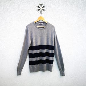 May include: A grey jumper with navy blue horizontal stripes. The long-sleeved crew neck jumper is hanging on a wooden hanger against a white wall. The Fila logo is visible on the upper left chest.