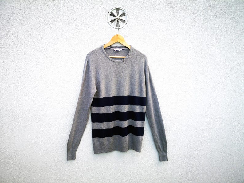 May include: A grey jumper with navy blue horizontal stripes. The long-sleeved crew neck jumper is hanging on a wooden hanger against a white wall. The Fila logo is visible on the upper left chest.