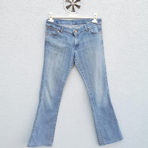 May include: Light blue denim jeans with a bootcut leg opening. The jeans are hanging on a white hanger against a white wall.