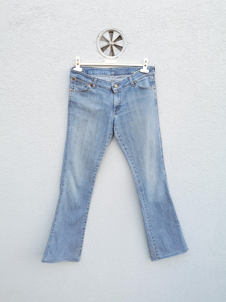 May include: Light blue denim jeans with a bootcut leg opening. The jeans are hanging on a white hanger against a white wall.