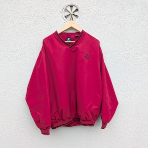 Può includere: Un antivento rosso con scollo a V e finiture nere, appeso a una gruccia di legno. La giacca presenta un piccolo logo sul petto, polsini ed orlo elasticizzati. Lo sfondo è un muro bianco strutturato.