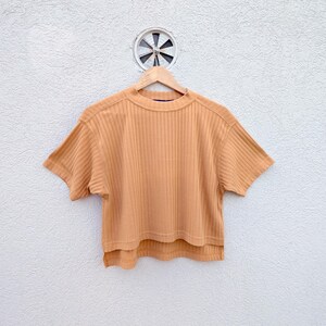 Puede incluir: Una camiseta de manga corta en tejido de punto acanalado de color tostado claro con cuello redondo. La prenda presenta una silueta cuadrada y corta con un dobladillo trasero ligeramente más largo, colgada en una percha.