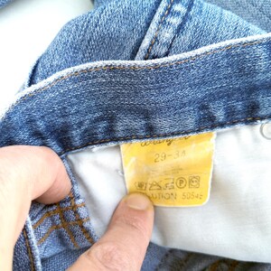 May include: Close-up of a blue denim jean waistband with a yellow Wrangler label that reads "29-34" and includes care instructions.