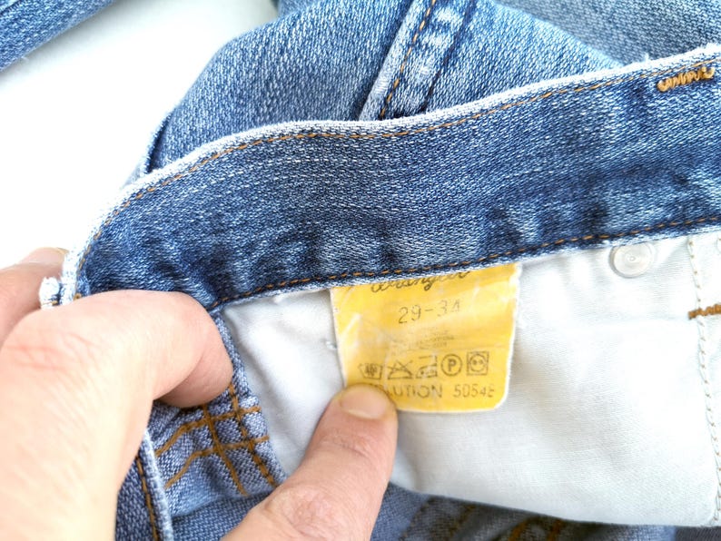 May include: Close-up of a blue denim jean waistband with a yellow Wrangler label that reads "29-34" and includes care instructions.