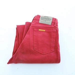 Puede incluir: Vaqueros de mezclilla rojos doblados con un bolsillo trasero y una pequeña etiqueta. Los vaqueros tienen un aspecto descolorido y una etiqueta rectangular que dice "JEANSEDWIN".