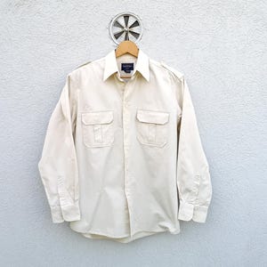 Peut inclure: Une chemise boutonnée à manches longues de couleur blanc cassé avec deux poches à rabat sur la poitrine. La chemise a un col et un devant boutonné.