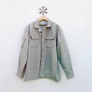 May include: A light olive green jacket with two chest pockets and two lower pockets. The jacket has long sleeves and a collared neckline. It is hanging on a wooden hanger against a white wall.