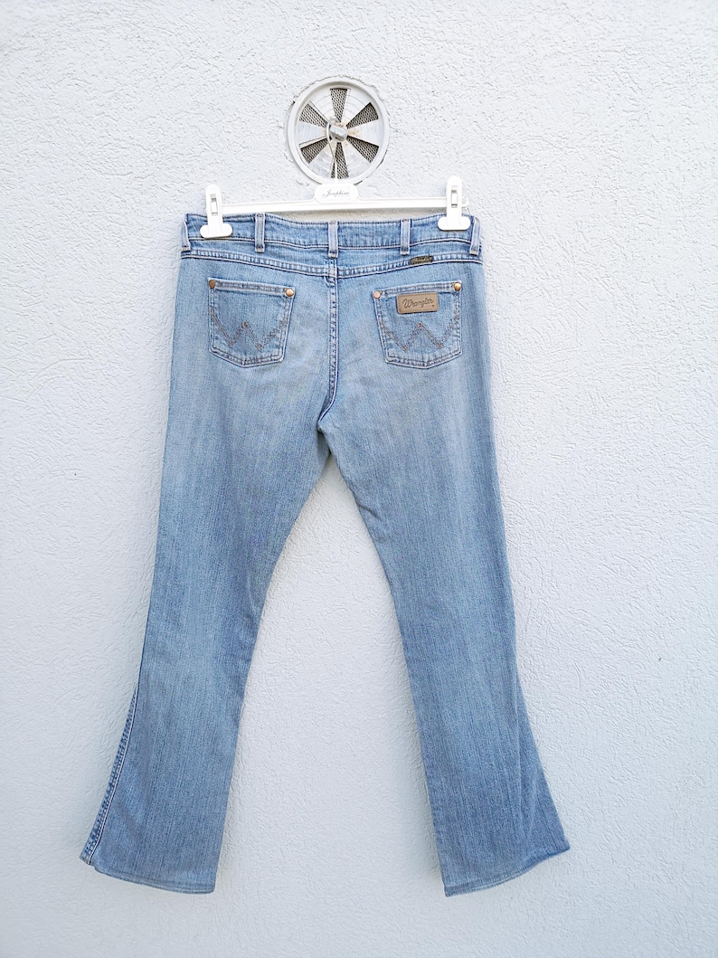 May include: A pair of light blue denim jeans with a bootcut leg. The jeans have a brown leather patch on the back with the Wrangler logo.