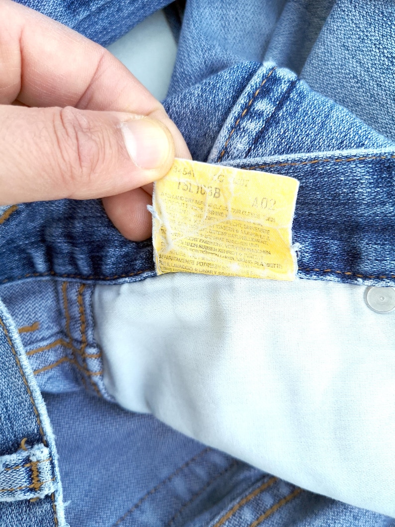 May include: A close-up of a blue denim jean pocket with a yellow care label that reads "15L 1038 40%".